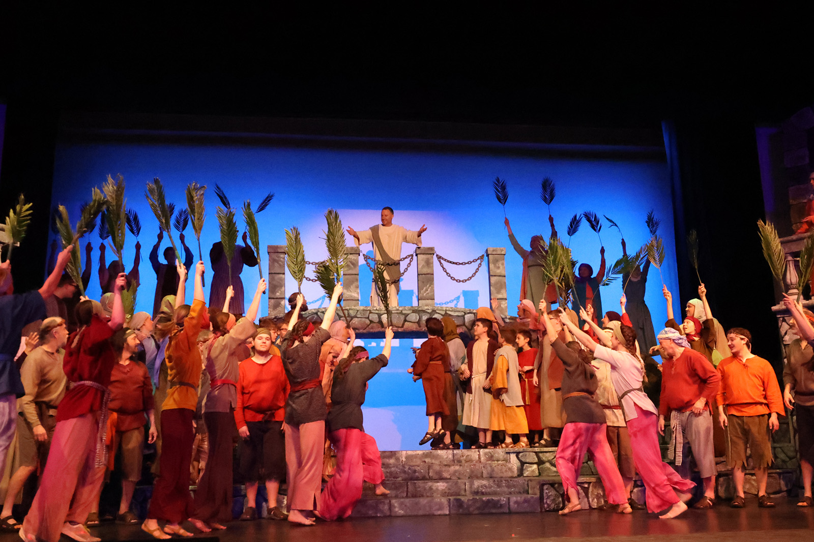 A crowd of exuberant people dressed in biblical attire hold palms as they greet Jesus.